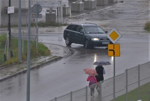 deszczowy dzień, kobieta i dziecko idą  chodnikiem z parasolami, jezdnią porusza się czarny samochód