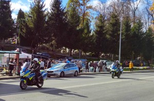 Dwa przejeżdżające motocykle policyjne i radiowóz oznakowany w rejonie cmentarza. W tle piesi i samochody.