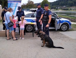 policjant z psem służbowym, obok policyjny radiowóz i stoisko profilaktyczne Policji - awatar