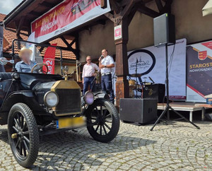 Przejeżdżający samochód retro, w tle na scenie policjant wygłaszający prelekcję oraz konferansjer.