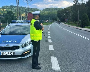 umundurowany policjant drogówki z laserowym miernikiem prędkości, obok radiowóz, w oddali jadące samochody