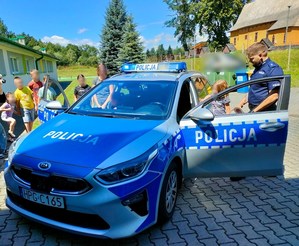 Bezpieczne Wakacje -umundurowany policjant prezentuje radiowóz oznakowany uczestnikom spotkania profilaktycznego