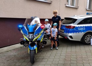 Bezpieczne Wakacje -wspólne zdjęcie policjantki i dzieci przy motocyklu służbowym, obok widoczny tył radiowozu oznakowanego.