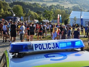 dach policyjnego radiowozu z lampą błyskową z napisem Policja, w tle uczestnicy Festiwalu Biegowego