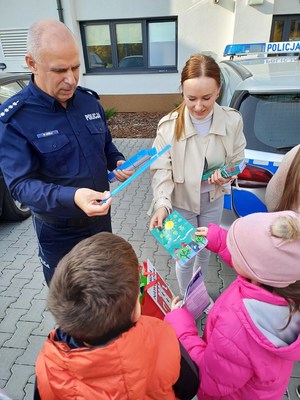 Funkcjonariusz oraz przedstawicielka gminy rozdają dzieciom pamiątkowe odblaski i ulotki