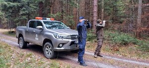 policjant i strażnik leśny patrzą przez lornetki, obok pojazd służbowy Straży Leśnej