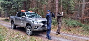 policjant i strażnik leśny patrzą przez lornetki, obok pojazd służbowy Straży Leśnej