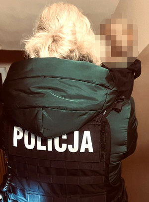 kobieta w zielonej kurtce i kamizelce z napisem Policja trzyma małego chłopca na rękach, stoją tłem, ich twarze są niewidoczne