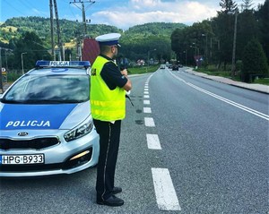 umundurowany policjant drogówki z laserowym miernikiem prędkości, obok radiowóz, w oddali jadące samochody