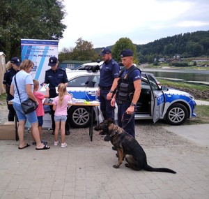 policjant z psem służbowym, obok policyjny radiowóz i stoisko profilaktyczne Policji