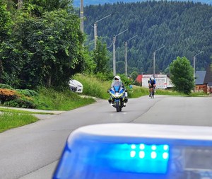 na pierwszym planie fragment lampy na dachu radiowozu, w oddali policjant na motocyklu służbowym, za nim kolarze