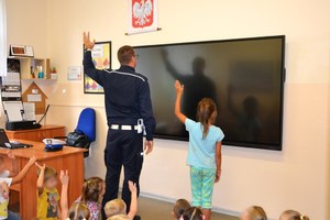 sala lekcyjna, policjant i uczennica podnoszą lewą dłoń, naśladują ich pozostali uczniowie