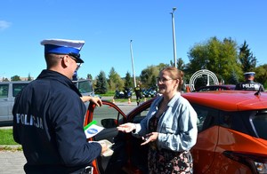 policjant drogówki wręcza kobiecie stojącej przy samochodzie odblask, ulotkę i naklejkę