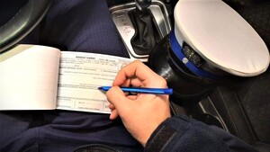 wnętrze radiowozu, policjant wypełnia bloczek mandatowy, obok policyjna czapka