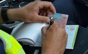 Policjant trzymając w ręku bloczek mandatowy oraz prawo jazdy wypisuje dokumenty.