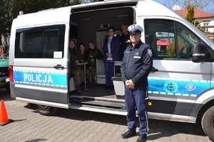 oznakowany radiowóz, policjant ruchu drogowego stoi obok, w środku dyrektor szkoły, policjantka prewencji oraz uczennice