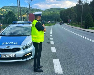 umundurowany policjant drogówki z laserowym miernikiem prędkości, obok radiowóz, w oddali jadące samochody