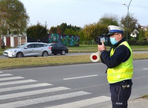 policjant z laserowym miernikiem prędkości, obok przejście dla pieszych, w tle jadące samochody