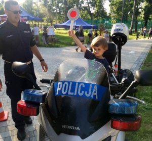 chłopiec siedzi na służbowym motocyklu i trzyma w ręce tarczę do zatrzymywania pojazdów, obok stoi policjant