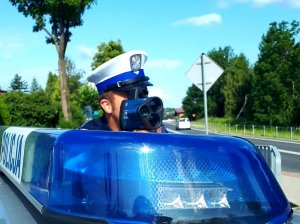 policjant z laserowym miernikiem prędkości, obok niebieska lampa na dachu radiowozu z napisem POLICJA