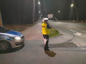 Policjant Wydziału Ruchu Drogowego z laserowym miernikiem prędkości, przód radiowozu oznakowanego