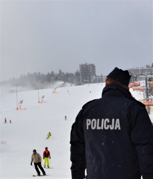 na pierwszym planie policjant prewencji odwrócony tyłem, w tle narciarze zjeżdżający ze stoku
