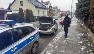 srebrny samochód z otwartą maską, przy nim umundurowany policjant, obok oznakowany radiowóz