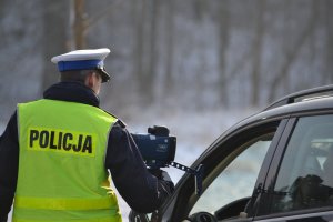 Policjant ruchu drogowego (biała czapka, żółta kamizelka z napisem Policja) podczas kontroli drogowej okazuje kierowcy zarejestrowany pomiar prędkości na ręcznym mierniku prędkości.