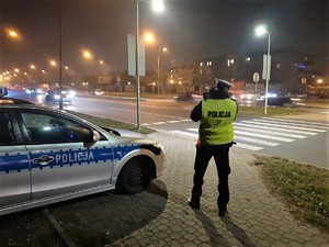 noc, policjant ruchu drogowego z laserowym miernikiem prędkości, obok zaparkowany oznakowany radiowóz