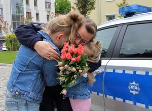 mama-policjantka w objęciach swoich córek z bukietem kwiatów w rękach, obok zaparkowany radiowóz