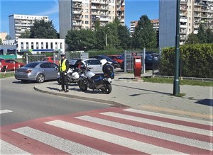 4. oznakowane przejście dla pieszych przy którym stoi umundurowany policjant  obok motocykl służbowy, w tle samochody i budynki