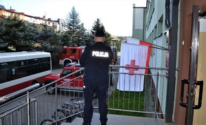 11. schody prowadzące do wejścia do szkoły, stojący policjant odwrócony tyłem, obok polskiej flagi, biała flaga z czerwonym krzyżem