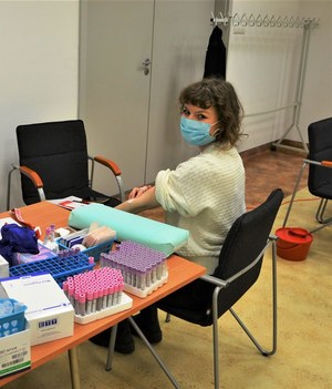 1. kobieta w maseczce siedzi przy stole z probówkami, uciskając miejsce po pobraniu krwi do badania