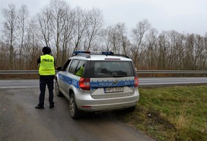 1. policjant w kamizelce odblaskowej z napisem policja oraz kasku ochronnym na głowie stoi przy drodze obok oznakowanego radiowozu