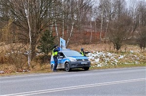 4. dwóch policjantów przy radiowozie, jeden z nich patrzy przez lornetkę, drugi unosi tarczę do zatrzymywania pojazdów