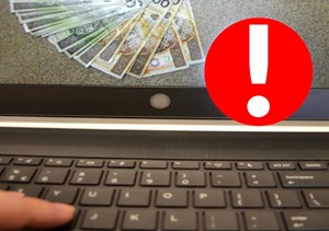 cyberoszustwa - klawiatura laptopa, na ekranie widoczne pieniądze, obok czerwona znak z wykrzyknikiem