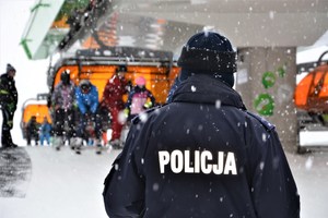 policjant odwrócony tyłem, na kurtce widoczny napis POLICJA, w tle narciarze wjeżdżający na górę wyciągiem