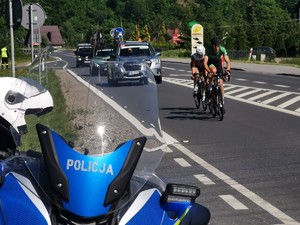 1. kolarze na trasie, za nimi samochody organizatora, na pierwszym planie fragment policyjnego motocykla