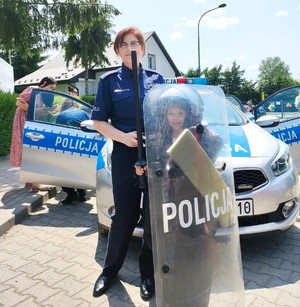 policjantka z dzieckiem ubranym w kask ochronny i tarczę, w tle radiowóz