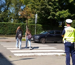dwie uczennice z plecakami przechodzą przez przejście dla pieszych, na chodniku stoi policjant drogówki