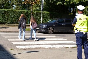 dwie uczennice z plecakami przechodzą przez przejście dla pieszych, na chodniku stoi policjant drogówki