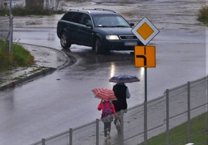 deszczowy dzień, kobieta i dziecko idą  chodnikiem z parasolami, jezdnią porusza się czarny samochód